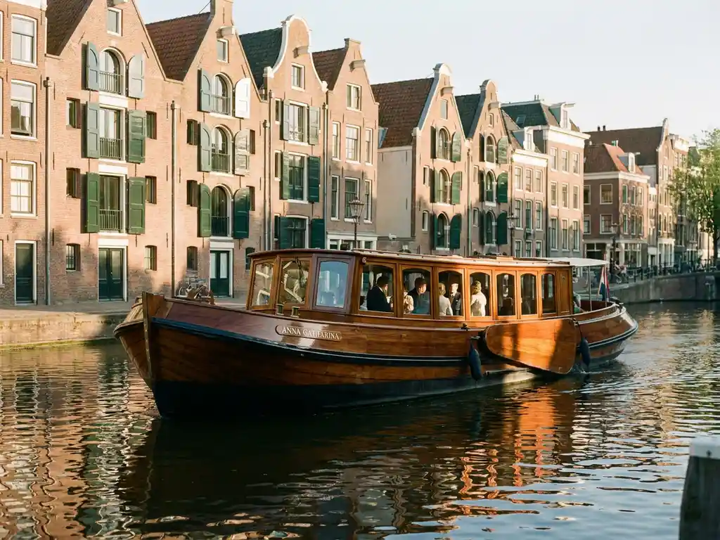 Klassieke salon tjalk uit 1910 vaart door Amsterdamse grachten, gepolijst irokohout glanst in warm middaglicht, historische gevels weerspiegeld in het water.