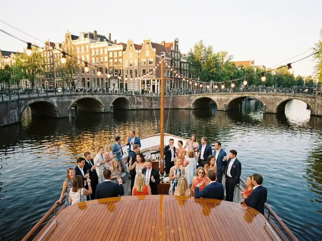 Luxe rondvaartboot vol zomerse gasten op Amsterdamse gracht, proosten bij gouden avondlicht met historische grachtenpanden op achtergrond.